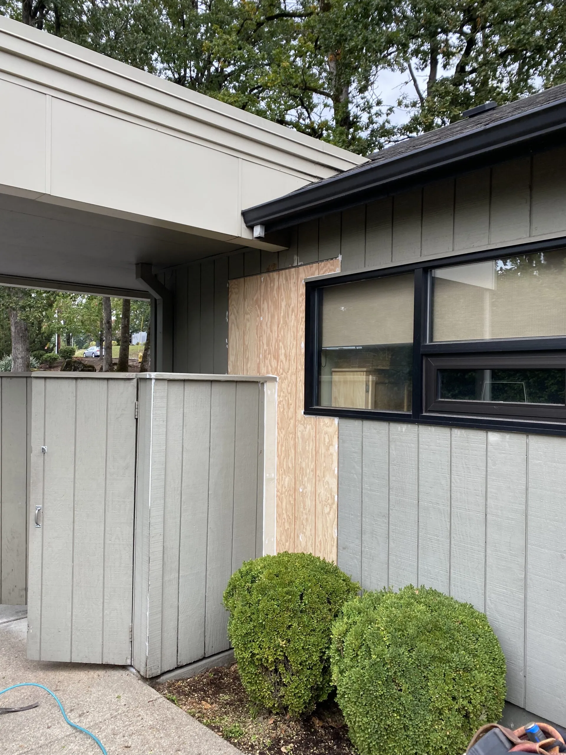 Modern home exterior featuring light gray vertical metal siding, black-framed windows, dark roof overhang, metal gate with