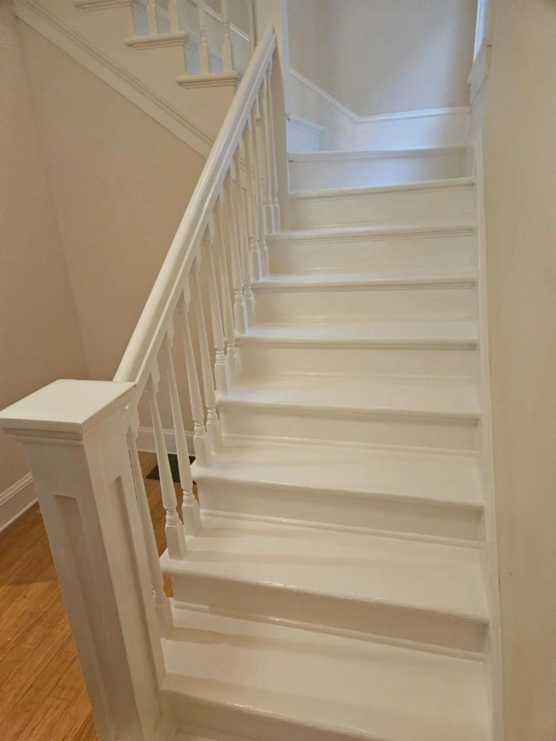 White painted staircase with white spindle railings descending to wood floor, light-filled stairwell with white walls.