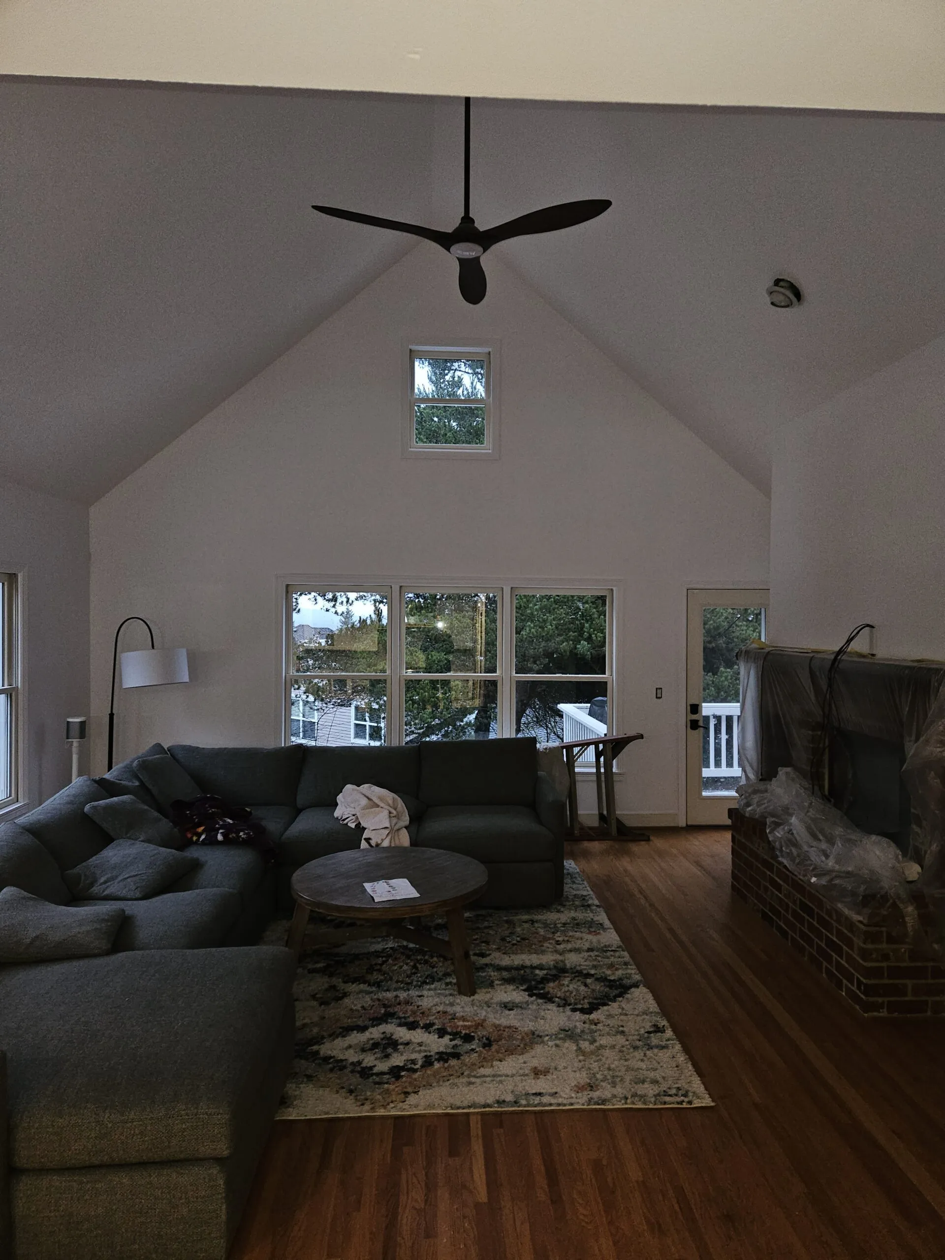 Bright living room with cathedral ceiling, black ceiling fan, large multi-pane windows overlooking trees, black sectional