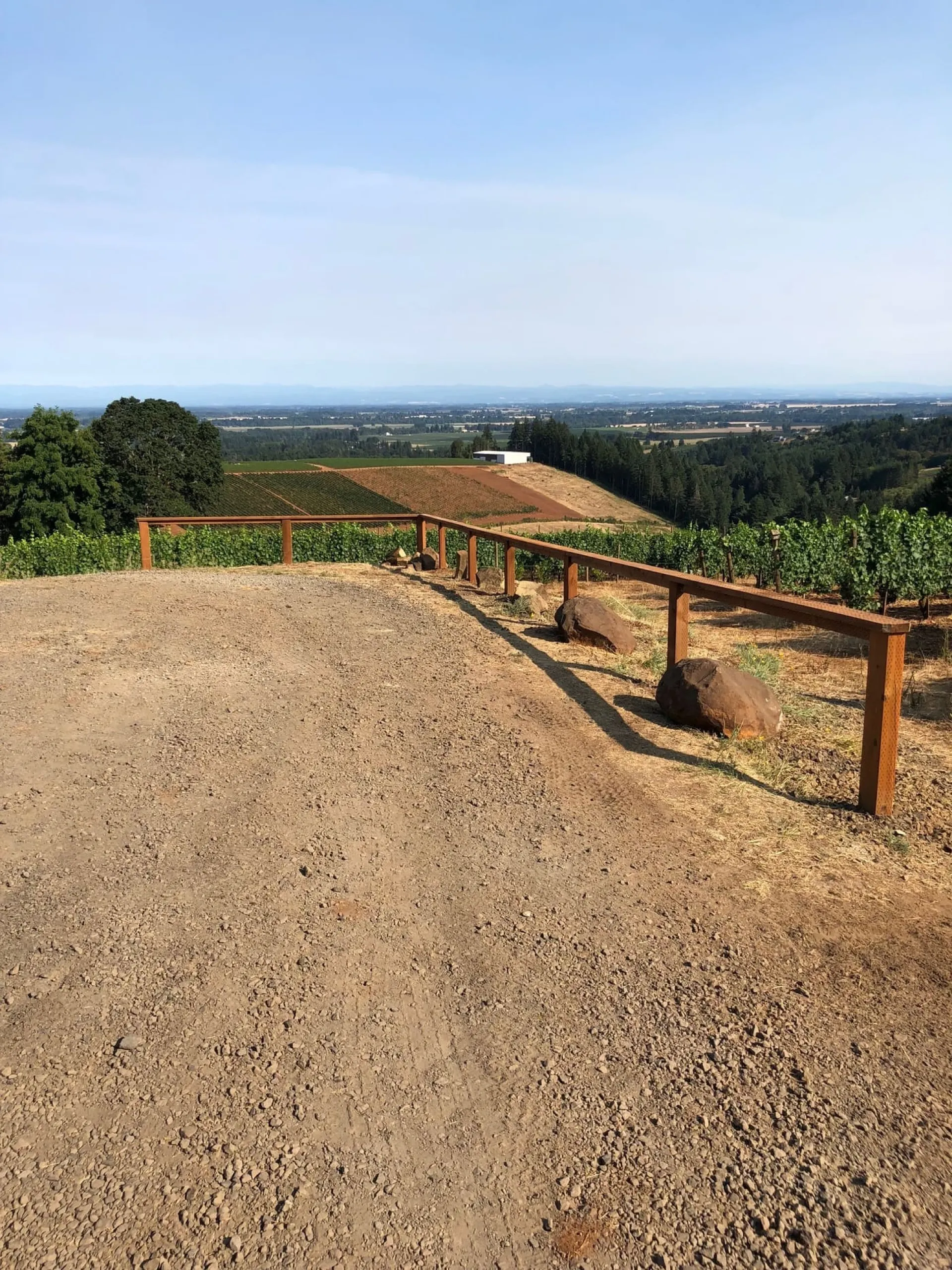 Wooden post-and-rail ranch fencing runs along a graded dirt driveway with forested hills and agricultural land visible in