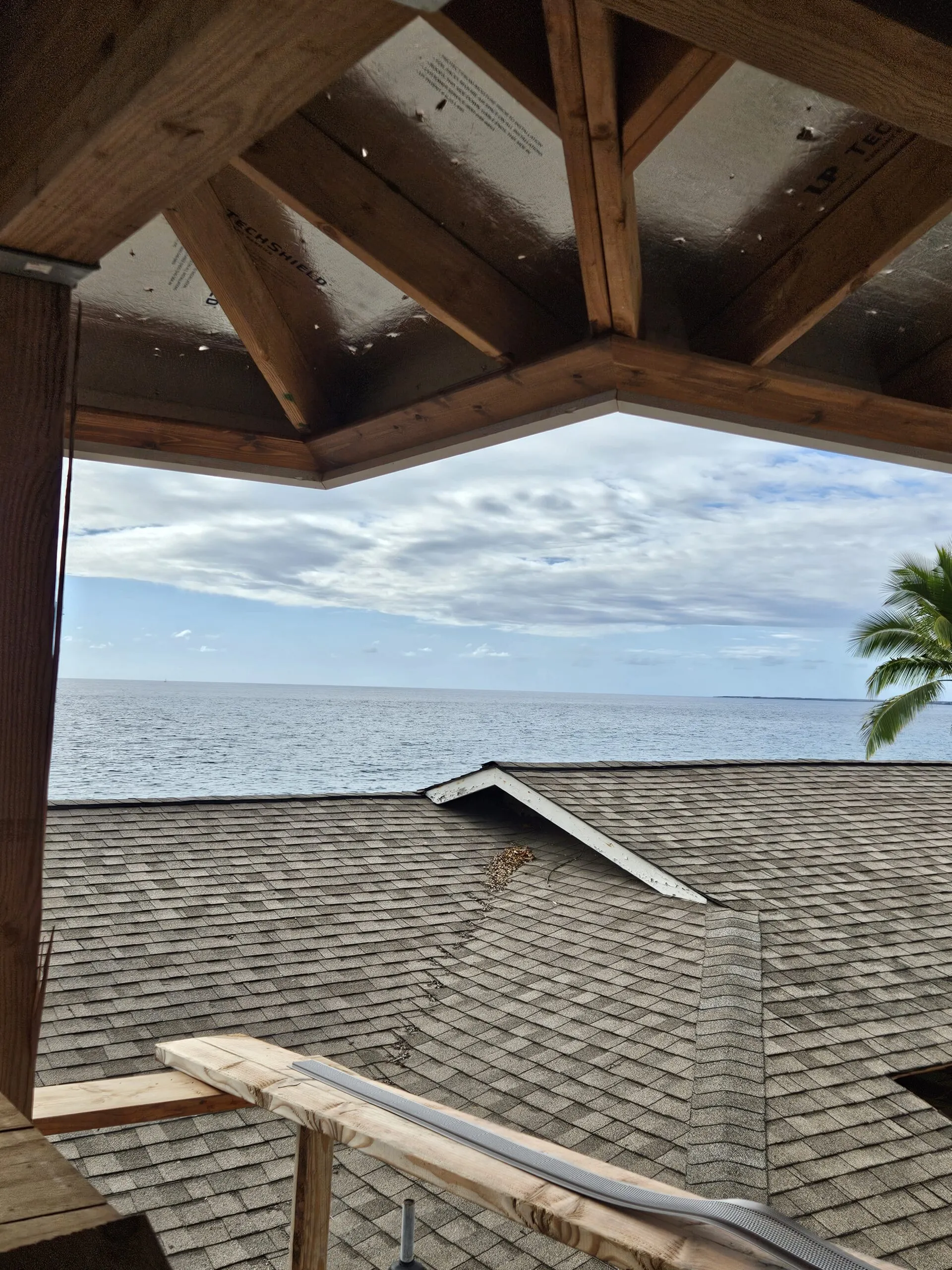 Newly shingled roof slope with ocean and sky visible beyond, framed by wooden beam structure and soffit.
