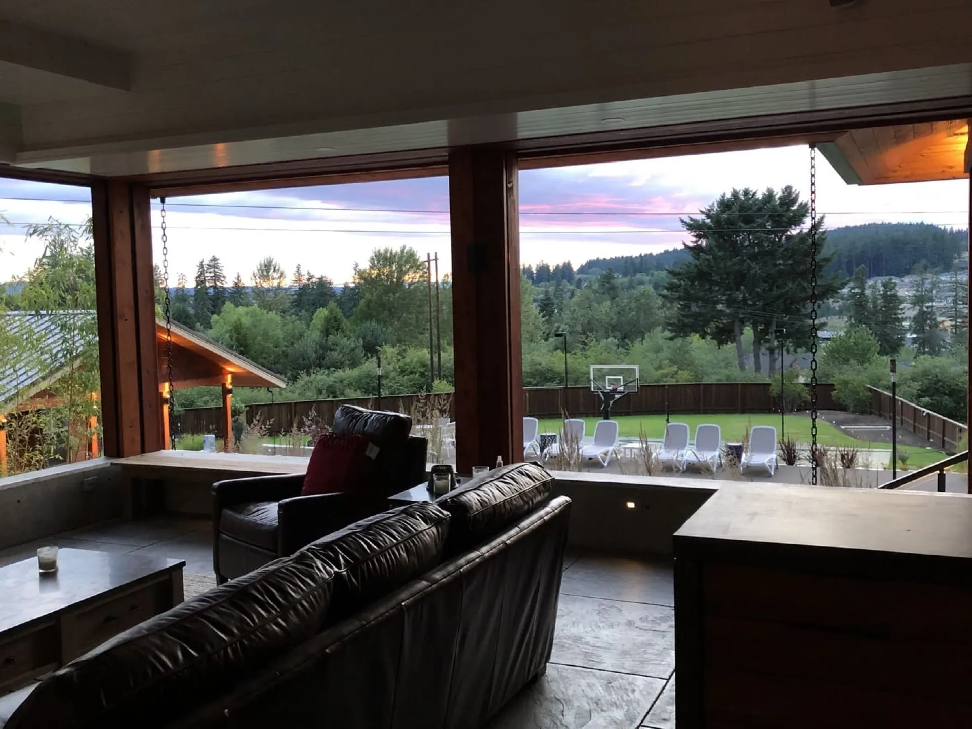 Modern living room with black leather sectional sofa facing large multi-panel windows, exterior view of forest, mountains