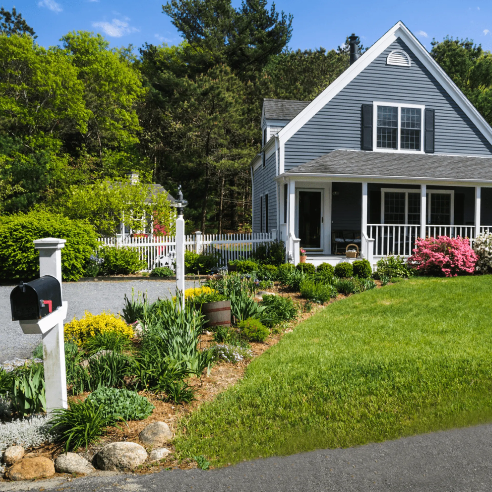 White farmhouse with gabled roof, front porch, mailbox, flowering shrubs, and sloped lawn with landscaped beds.